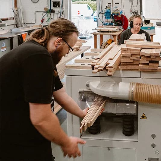 Een medewerker voert houten latten door de schaafmachine; een collega werkt op de achtergrond.