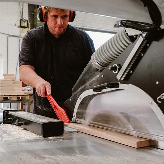 Een medewerker met gehoorbescherming zaagt een eiken plank op de zaagtafel.