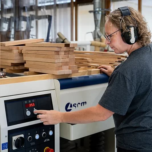 Een medewerker met gehoorbescherming bedient een bewerkingsmachine, naast een stapel houten latten.