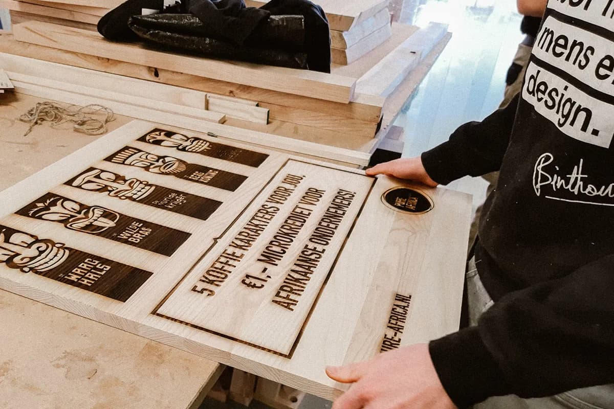 Een medewerker van Binthout houdt een lasergegraveerde houten plank met eigen ontwerp vast, naast een stapel hout.