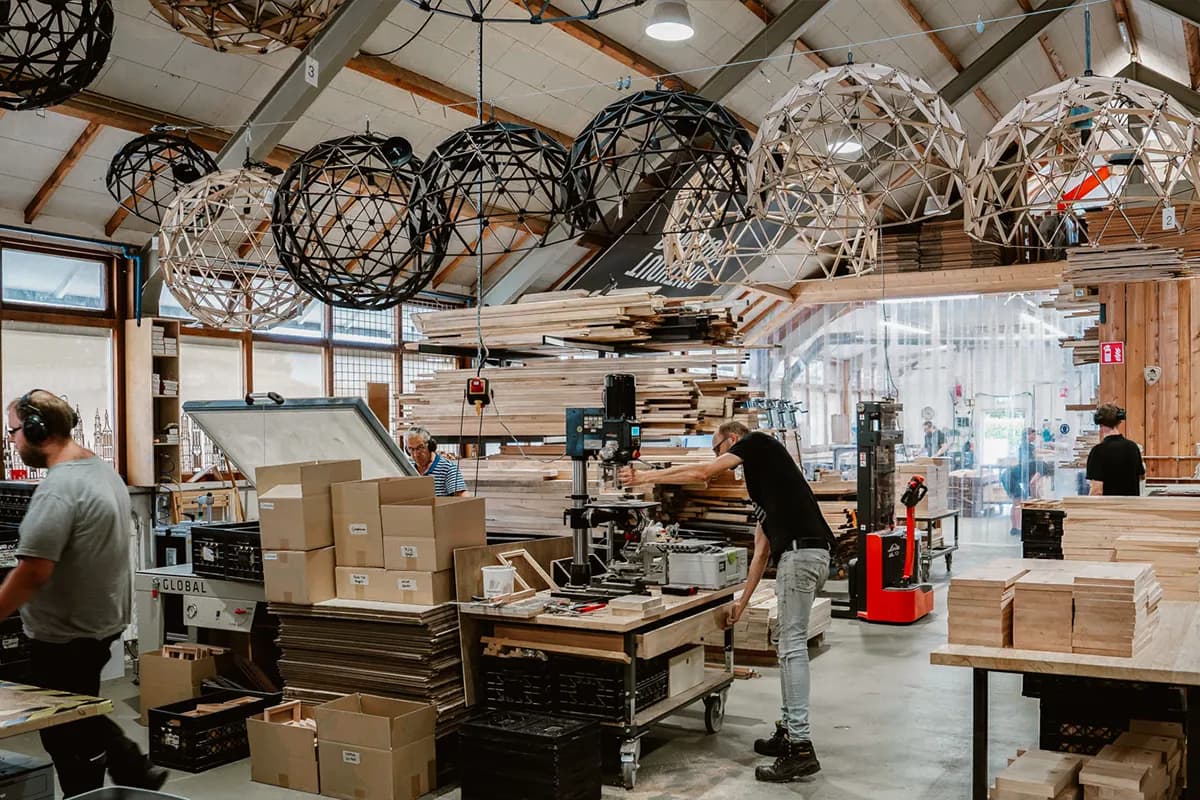 De werkplaats van Binthout: medewerkers aan het werk tussen stapels hout, onder houten hanglampen.